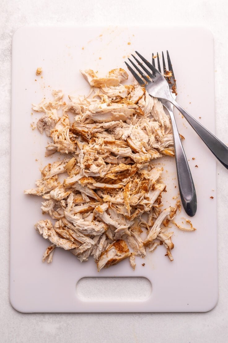 planche à découper blanche avec du poulet effiloché et deux fourchettes.