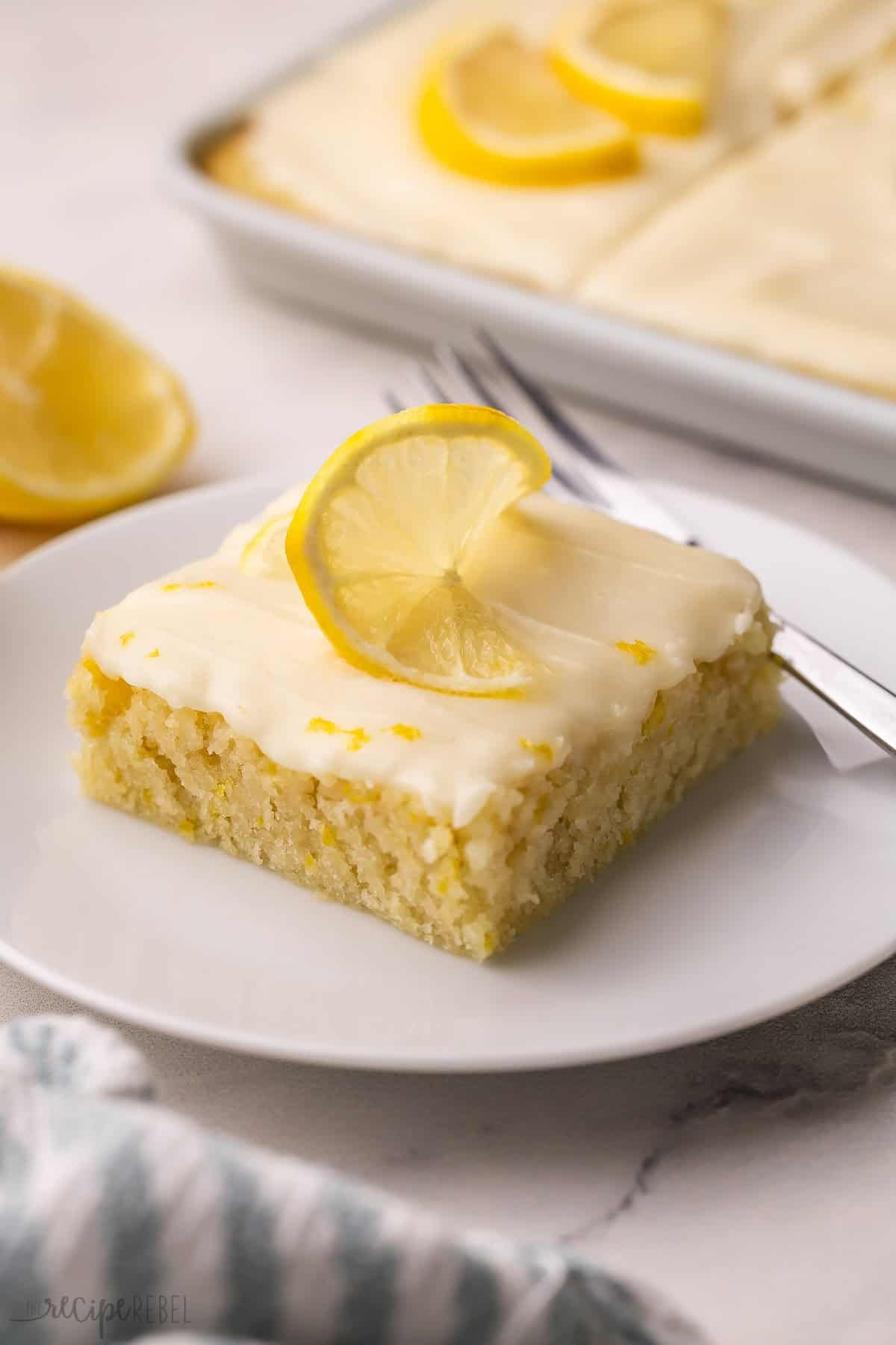 Ce gâteau au citron moelleux est infusé de saveurs vives et citronnées grâce à du jus de citron frais et du zeste. Prêt en moins de 40 minutes et surmonté d'un glaçage au citron pétillant, c'est l'équilibre parfait entre douceur et acidité ! un morceau de gâteau au citron sur une assiette blanche avec une fine tranche de citron sur le dessus.