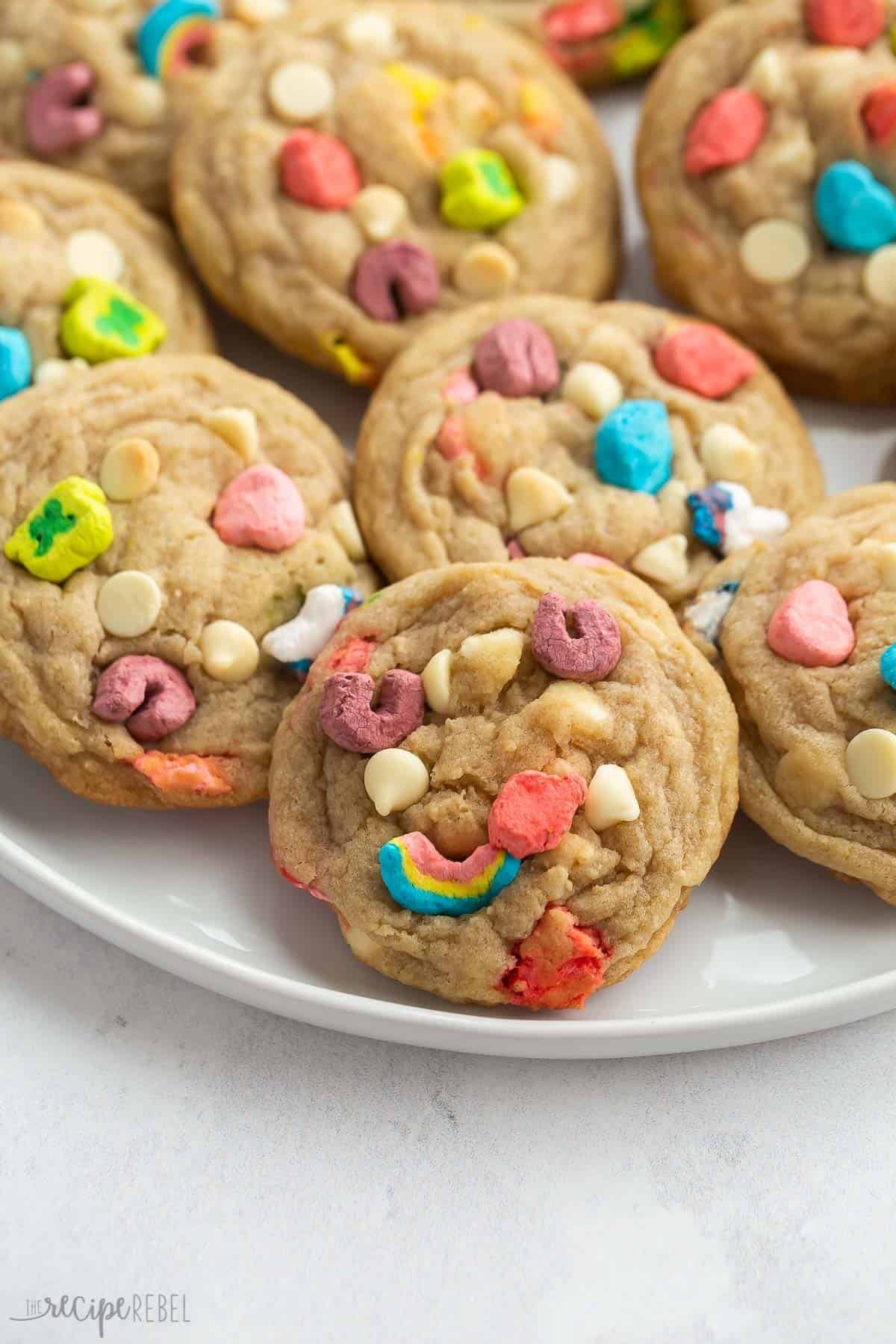 Ces Cookies aux Lucky Charms et chocolat blanc sont parfaitement moelleux et préparés avec des céréales Lucky Charms et des guimauves. Idéaux pour la Saint-Patrick ! Comprend une vidéo de recette étape par étape. gros plan sur des cookies aux lucky charms sur une assiette blanche.