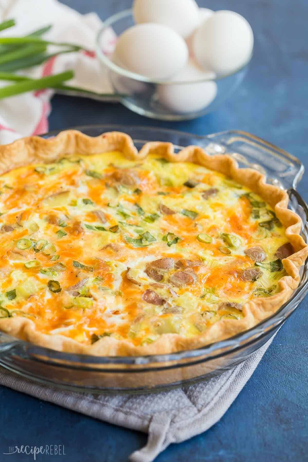Cette quiche aux pommes de terre chargée est facile à préparer et pleine de saveurs ! C'est la recette de quiche parfaite pour les fêtes ou un dîner en semaine. gros plan de quiche aux pommes de terre chargée dans un plat à tarte sur fond bleu foncé.