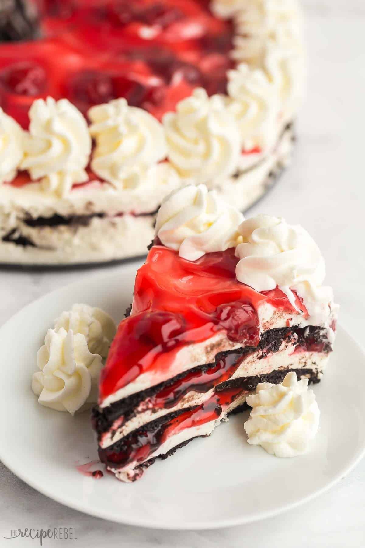 Ce gâteau glacé sans cuisson aux cerises noires est un dessert facile qui est parfait pour l'été ! Il ne nécessite que QUATRE ingrédients. Un morceau de gâteau glacé chocolat cerise sur une assiette blanche avec de la crème fouettée.