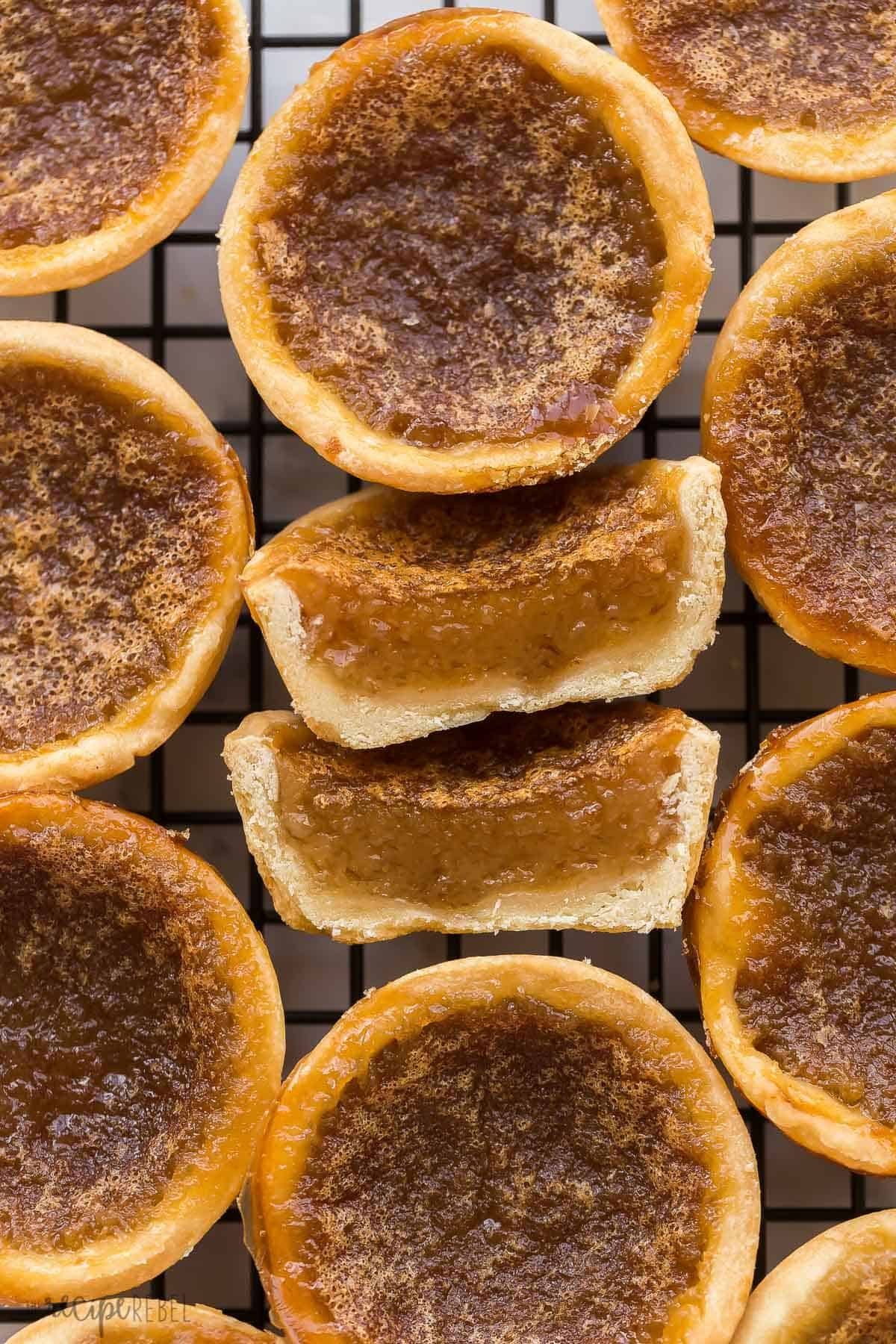 Ces Tartelettes au Beurre sont un classique canadien qui se prépare en seulement 25 minutes ! Des coques de pâtisserie maison sont remplies d'une garniture riche, au sucre brun (raisins en option !), puis cuites jusqu'à ce qu'elles soient beurrées et feuilletées à l'intérieur et gooey à l'intérieur. image en gros plan d'une tartelette au beurre coupée en deux sur un grille