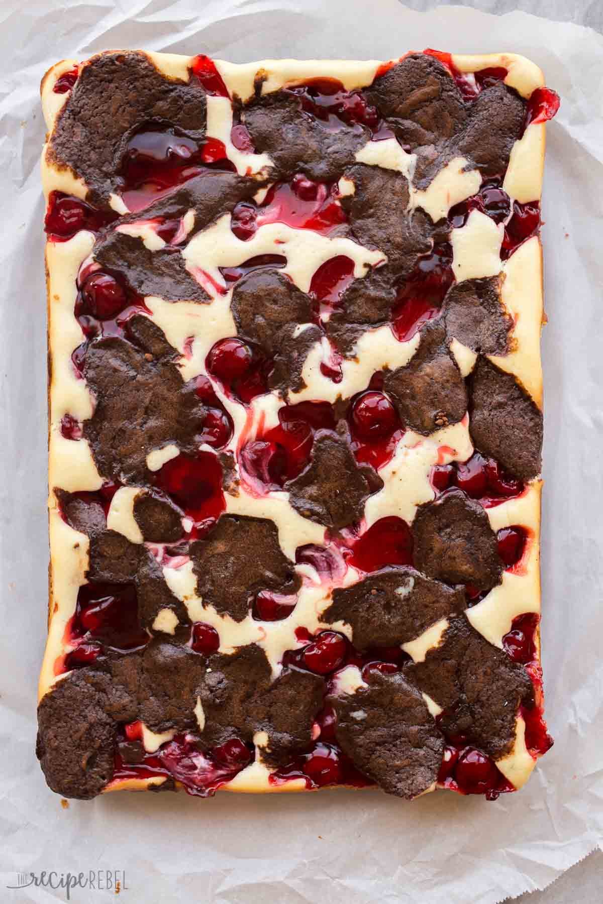 Plaque entière de brownies au cheesecake aux cerises non coupés dans un moule tapissé de papier sulfurisé.