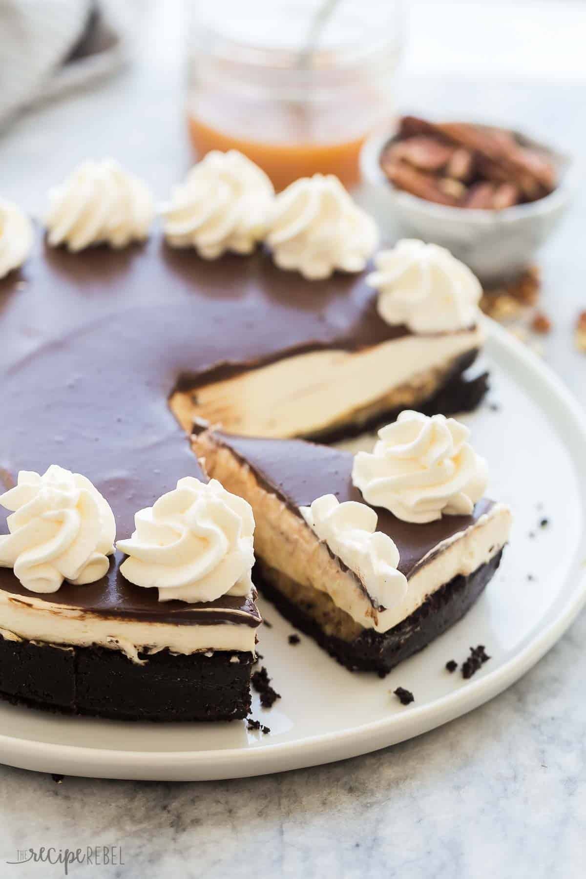 Cheesecake aux tortues sans cuisson a une croûte de biscuits Oreo, une couche de caramel aux noix de pécan faite maison, une couche de cheesecake crémeux et une ganache au chocolat riche. Prêt à l'avance ! Cheesecake aux tortues entier sans cuisson, garni de ganache, de crème fouettée et de caramel.