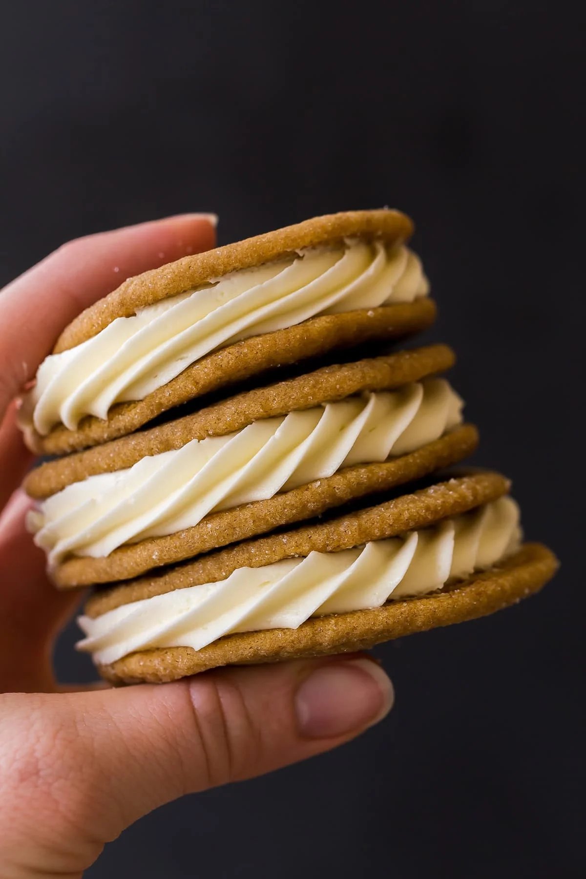 Les Cookies Sandwichs au Chocolat Blanc et aux Épices sont moelleux, fondants et garnis d'un glaçage au chocolat blanc fait maison. Ils sont légèrement épicés, avec un enrobage de sucre croquant et une garniture crémeuse. Le cookie de Noël parfait, facile à congeler ! #cookies #dessert #Noël Une main tenant une pile de cookies sandwich aux épices sur fond sombre.