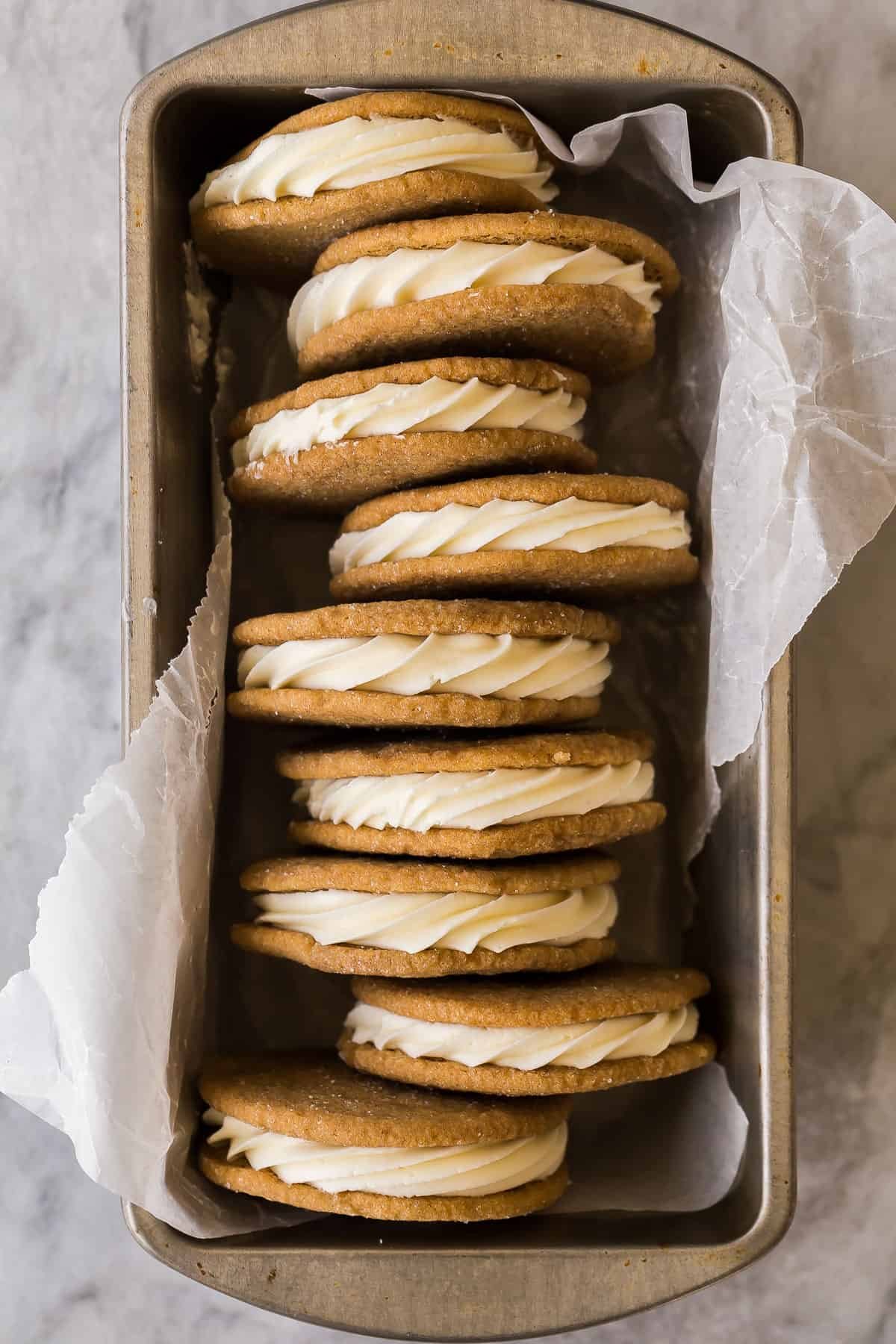 Les Cookies Sandwichs au Chocolat Blanc et aux Épices sont moelleux, fondants et garnis d'un glaçage au chocolat blanc fait maison. Ils sont légèrement épicés, avec un enrobage de sucre croquant et une garniture crémeuse. Le cookie de Noël parfait, facile à congeler ! #cookies #dessert #Noël Des cookies sandwich au chocolat blanc empilés dans une boîte métallique.